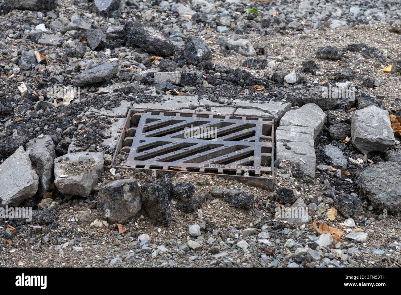 A rectangular cast iron storm drain grate sits surrounded by crumbling concrete kerbing, crushed gravel, and broken asphalt debris during urban road r Stock Photo