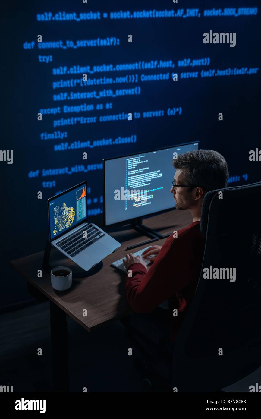 Software engineer sits at a desk with multiple monitors showing script and data science graphs in a dark room with programming code on background, works on AI and machine learning project. Copy space Stock Photo