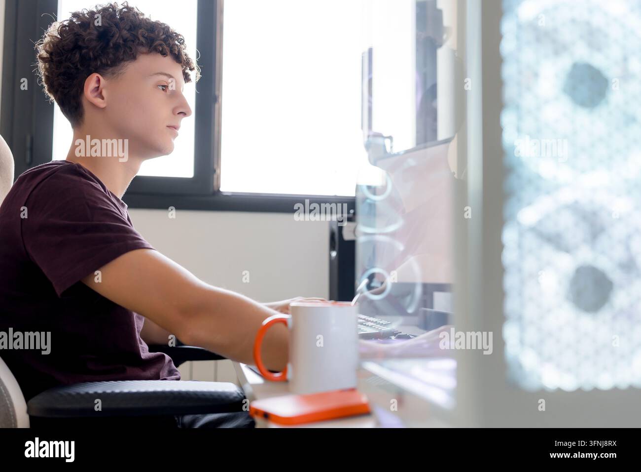 Young man works at a desktop gaming computer in a home office, using AI tools while seated at a desk beside a mug and smartphone in daylight. Stock Photo