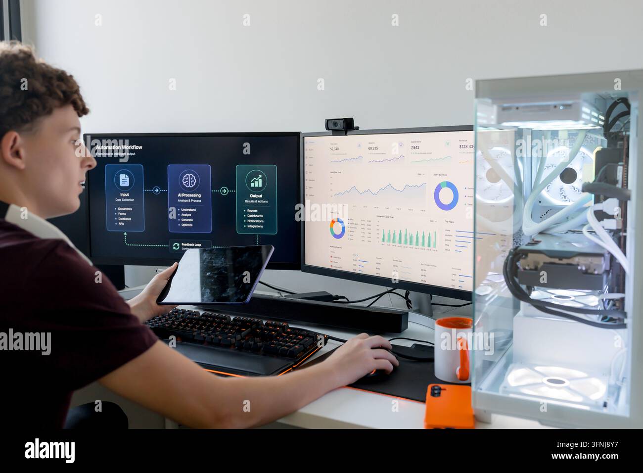 Young man works with AI tools and analytics dashboards on dual monitors, using a tablet beside a custom gaming computer in a modern home office. Stock Photo