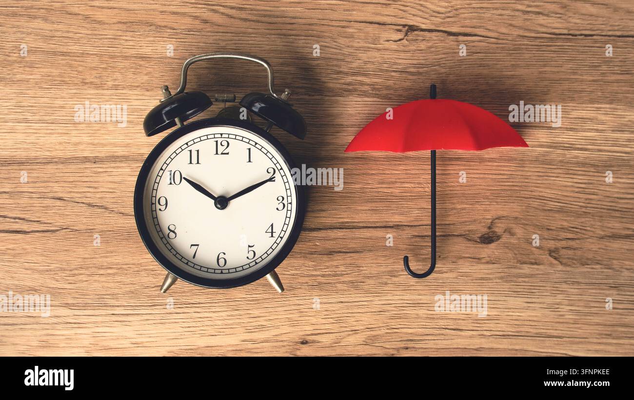 Black vintage alarm clock beside small red umbrella on wooden table time protection and life insurance concept top view. Stock Photo