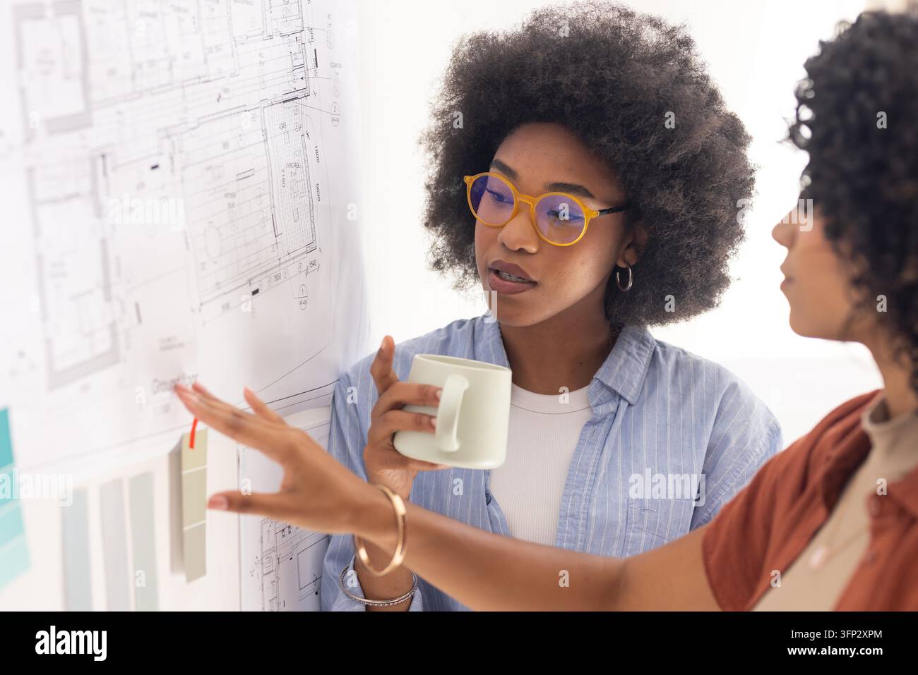 Diverse female designers studying pinned floor plans with color cards in studio, holding mug. Blueprint, architect, collaboration, brainstorming, workspace, modern, bright Stock Photo