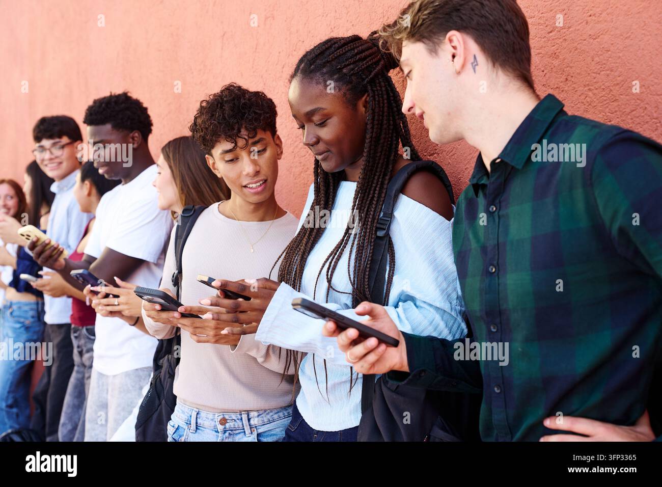 Group of multiethnic young students standing along a wall, using their smartphones to text and engage with social media Stock Photo