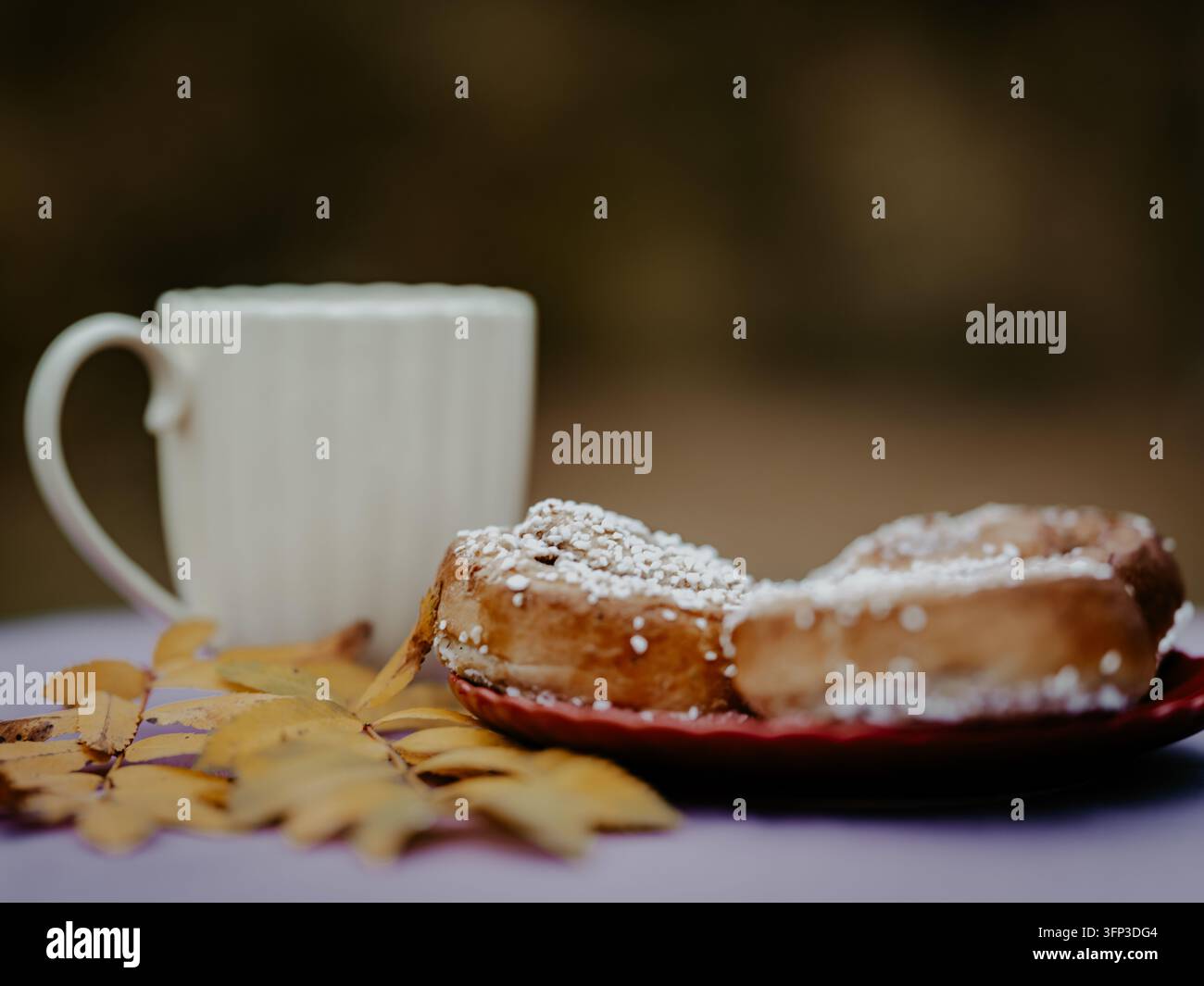 Swedish cinnamon buns topped with pearl sugar on a red plate, with a white coffee cup and yellow autumn leaves in the background. Stock Photo