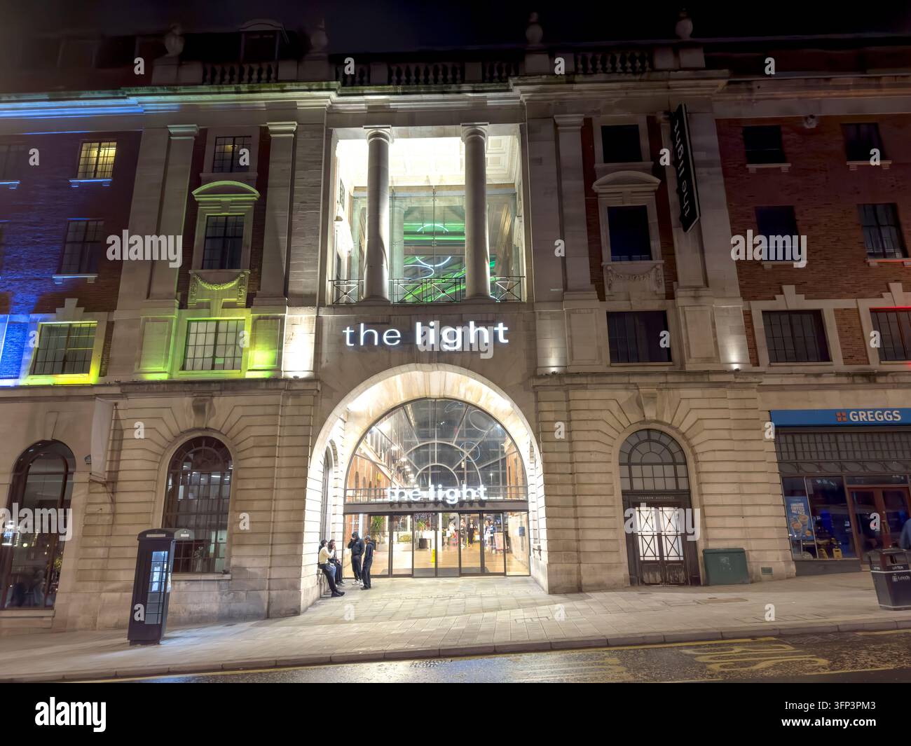The Light shopping and leisure complex entrance is illuminated after dark, framed by substantial stone architecture and pedestrians. Stock Photo