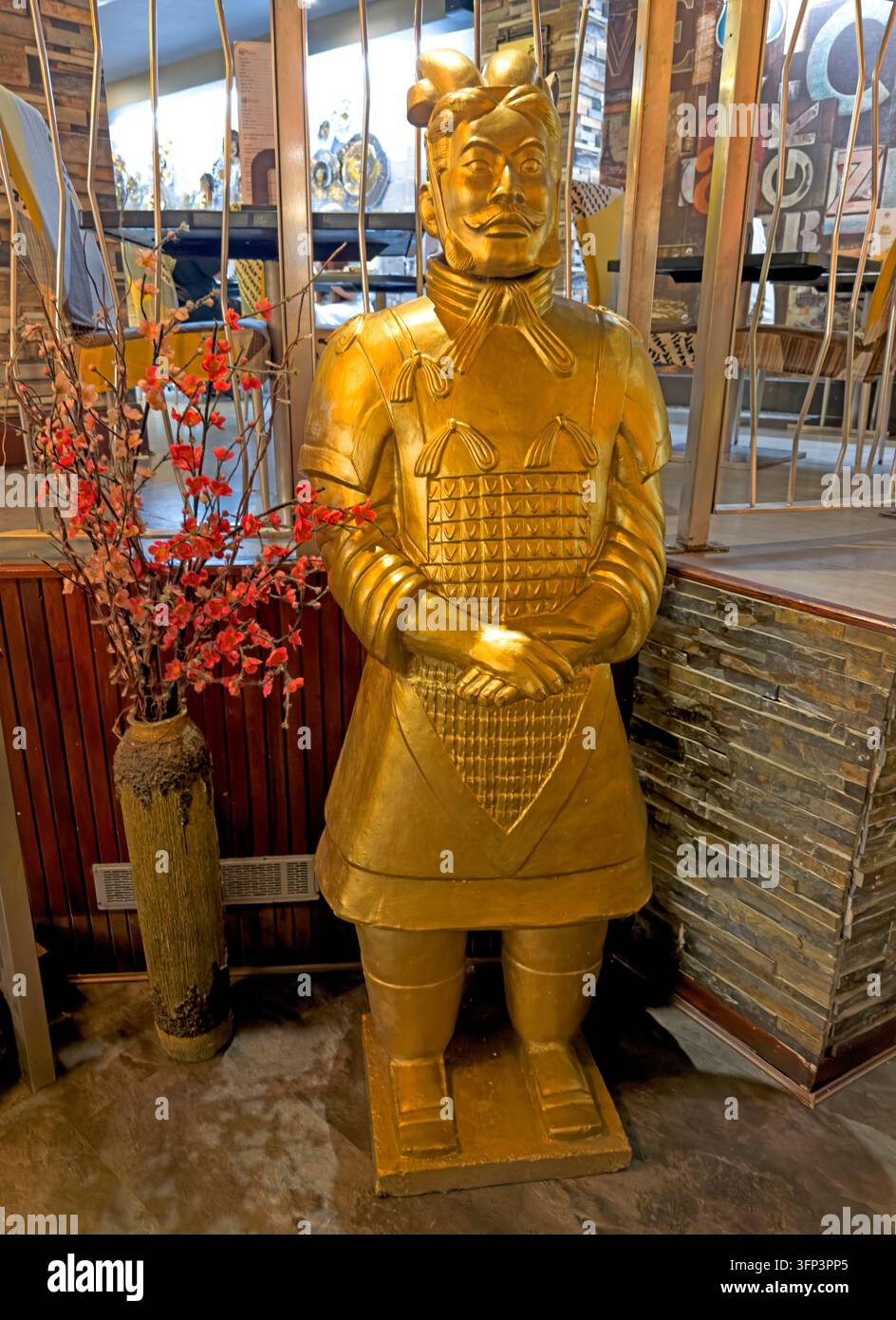 A gold-painted reproduction Chinese terracotta warrior stands inside a restaurant or hospitality interior, behind or beside decorative branches. Stock Photo