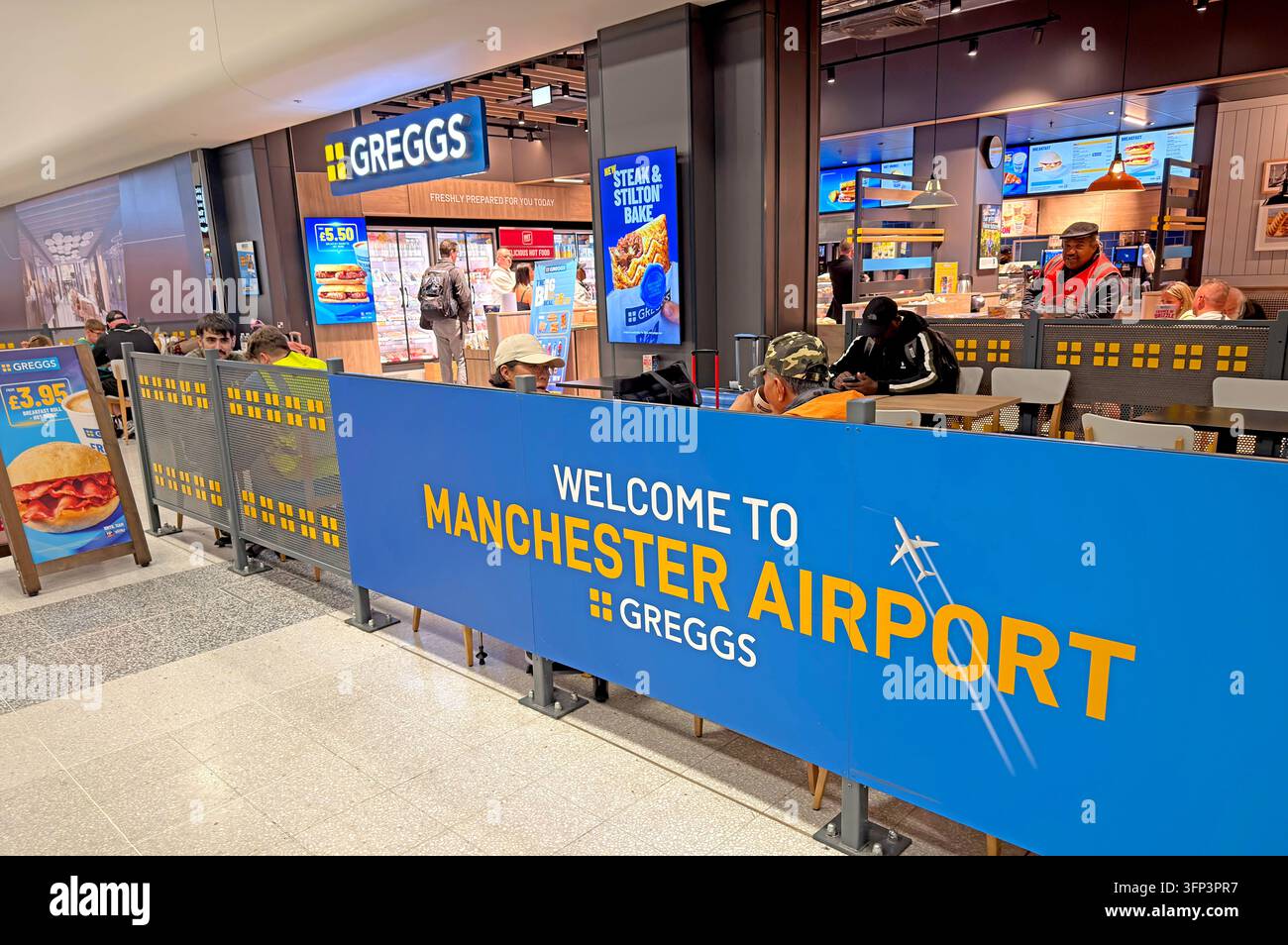 A Greggs bakery counter with bright signage and a blue Manchester Airport welcome barrier sits inside an airport terminal. Stock Photo