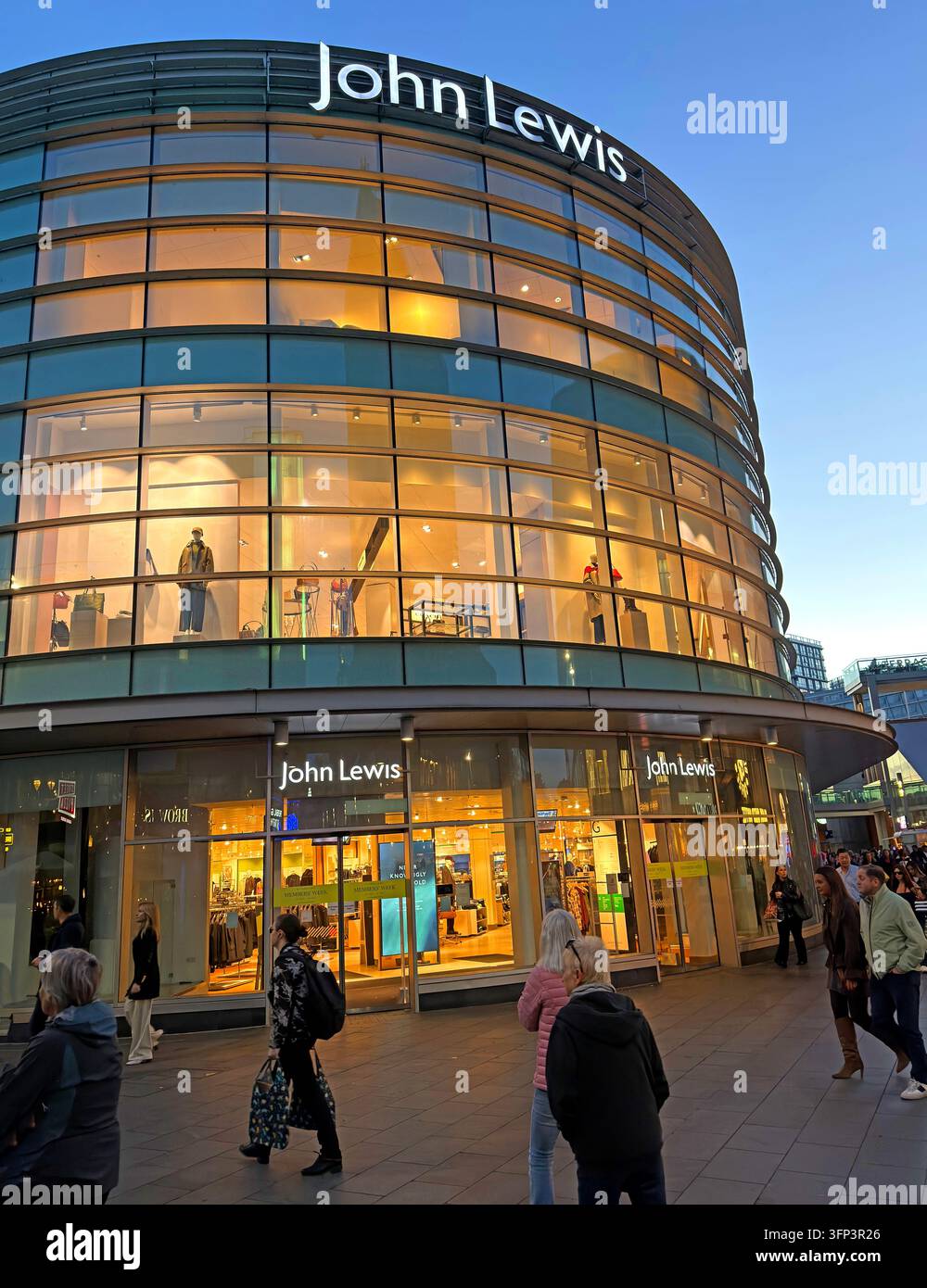 The curved glass façade of John Lewis is illuminated at dusk, with branding above ground-floor entrances and pedestrians nearby. Stock Photo