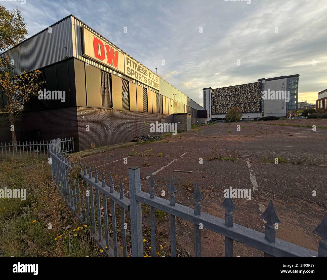 The exterior of a large former DW Sports and Fitness retail and gym building stands beside a vacant-looking forecourt and perimeter railings. Stock Photo