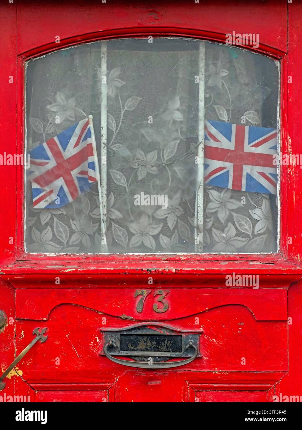 A red-painted entrance door numbered 73 has a glazed upper panel, patterned curtains, letterbox and two Union Jack flags displayed inside. Stock Photo