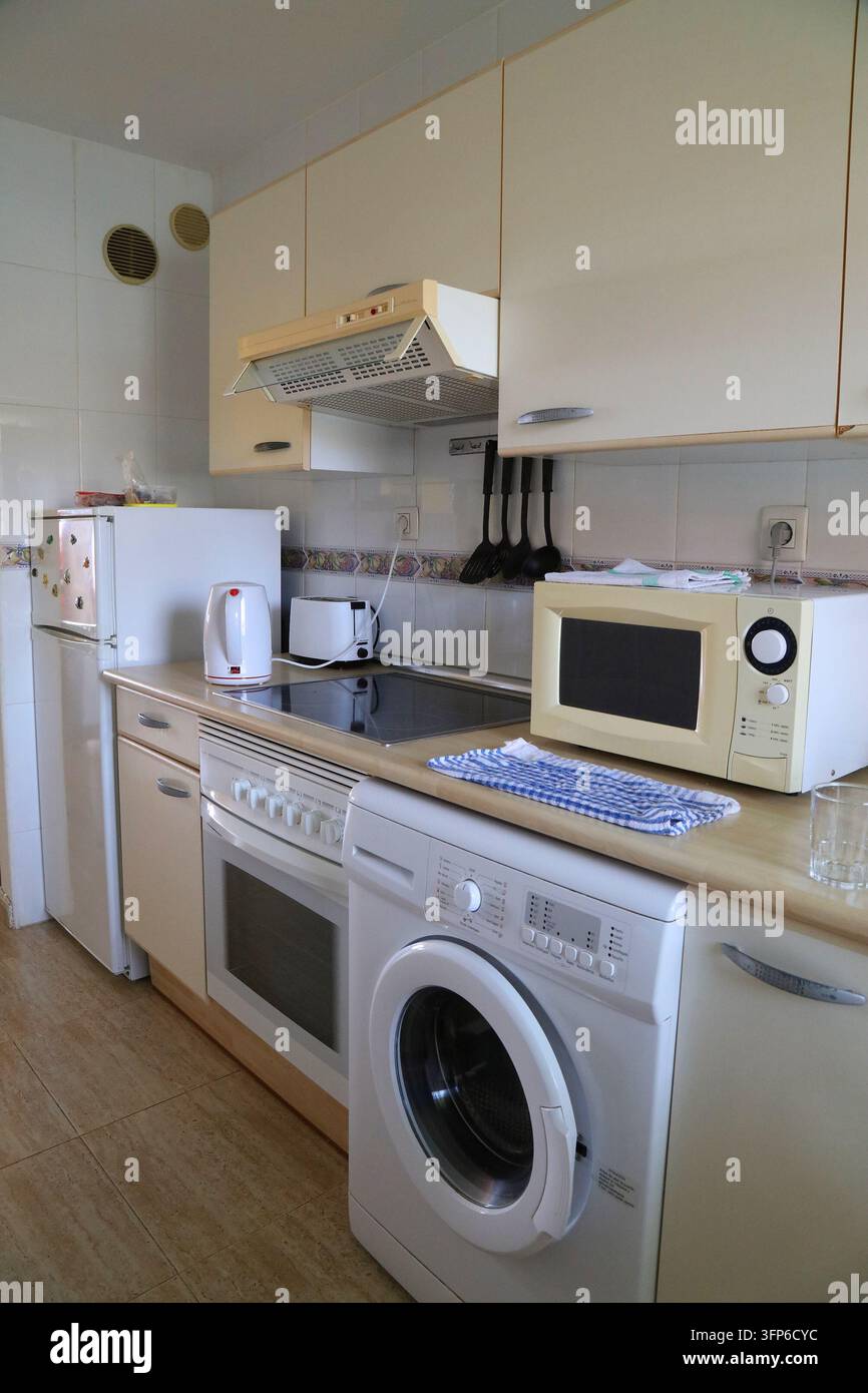 Generic basic normal kitchen interior in Spain. Kitchen with fridge, oven, microwave, washing machine and wall mounted overhead cabinets. Stock Photo