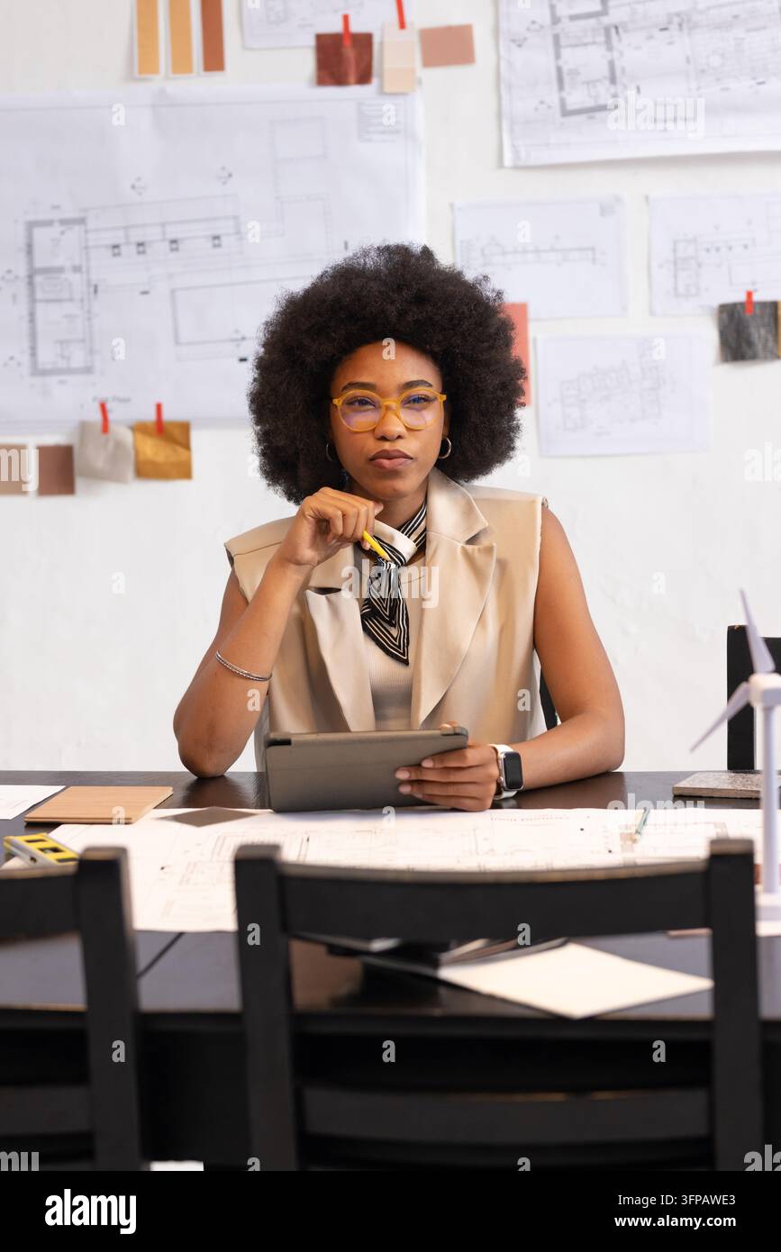 Prompted by drawings and swatches, woman sitting in studio wearing neck scarf reviewing tablet. Architect, designer, blueprint, materials, textures, workspace, office Stock Photo