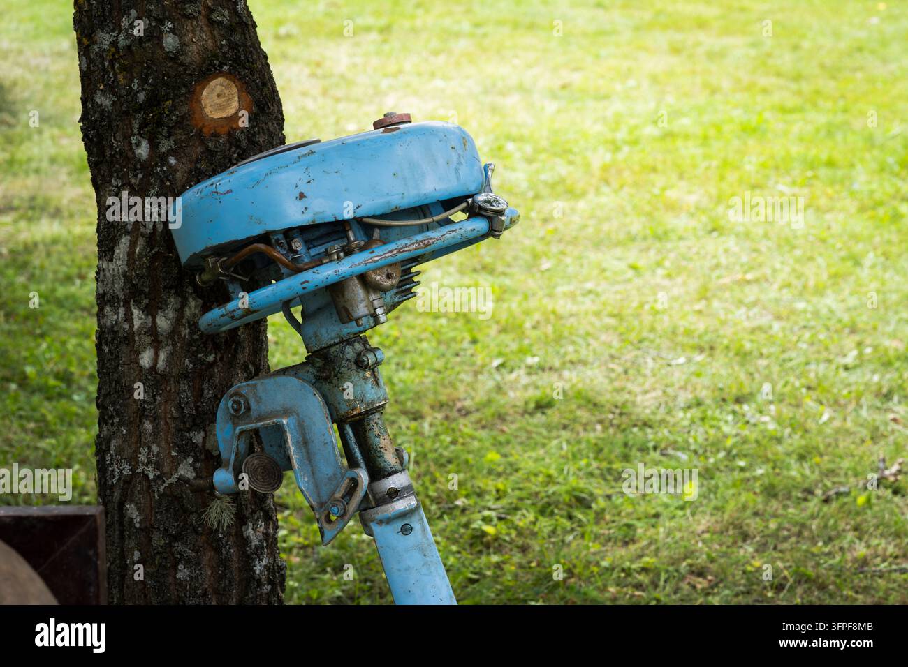 Vintage blue outboard boat motor with weathered paint, exposed mechanical components and signs of age, displayed beside a tree on a outdoor market. Stock Photo