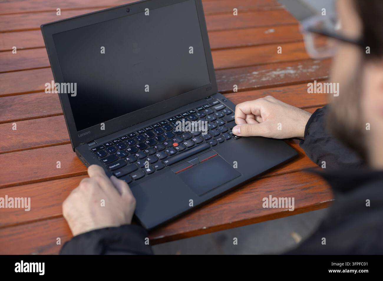 A partially visible person uses an open laptop on a wooden outdoor table. The close overhead view shows hands, keyboard, blank screen, and shallow bac Stock Photo