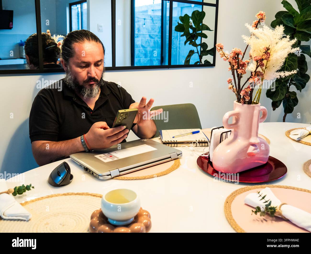 Person using a smartphone while seated at a dining table with a laptop ...