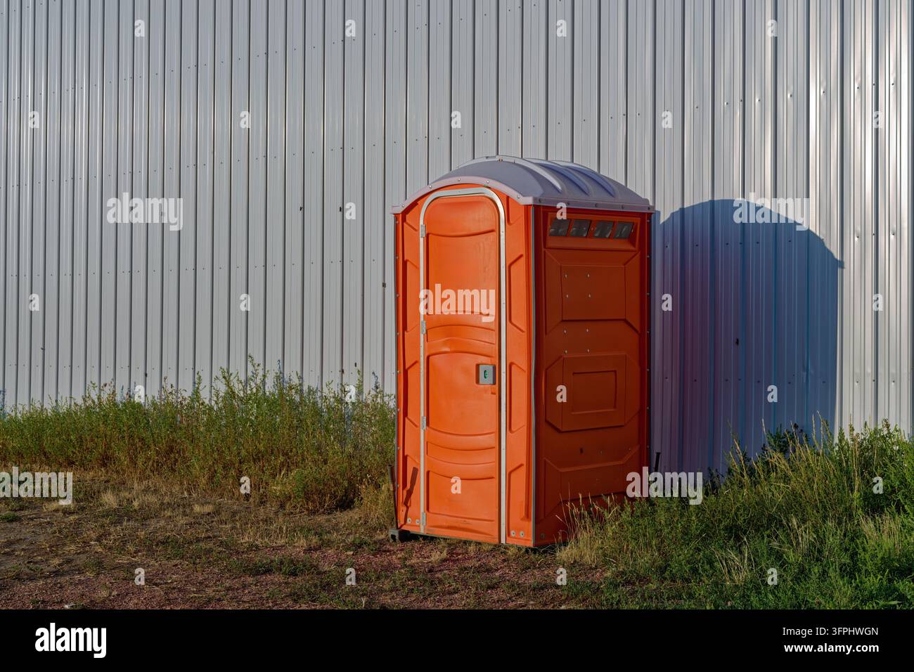 An orange outdoor toilet standing next to a corrugated metal wall as ...