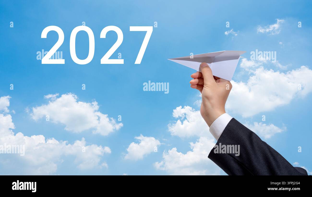 Businessman hand launching a paper airplane towards 2027 text in a blue sky. Stock Photo