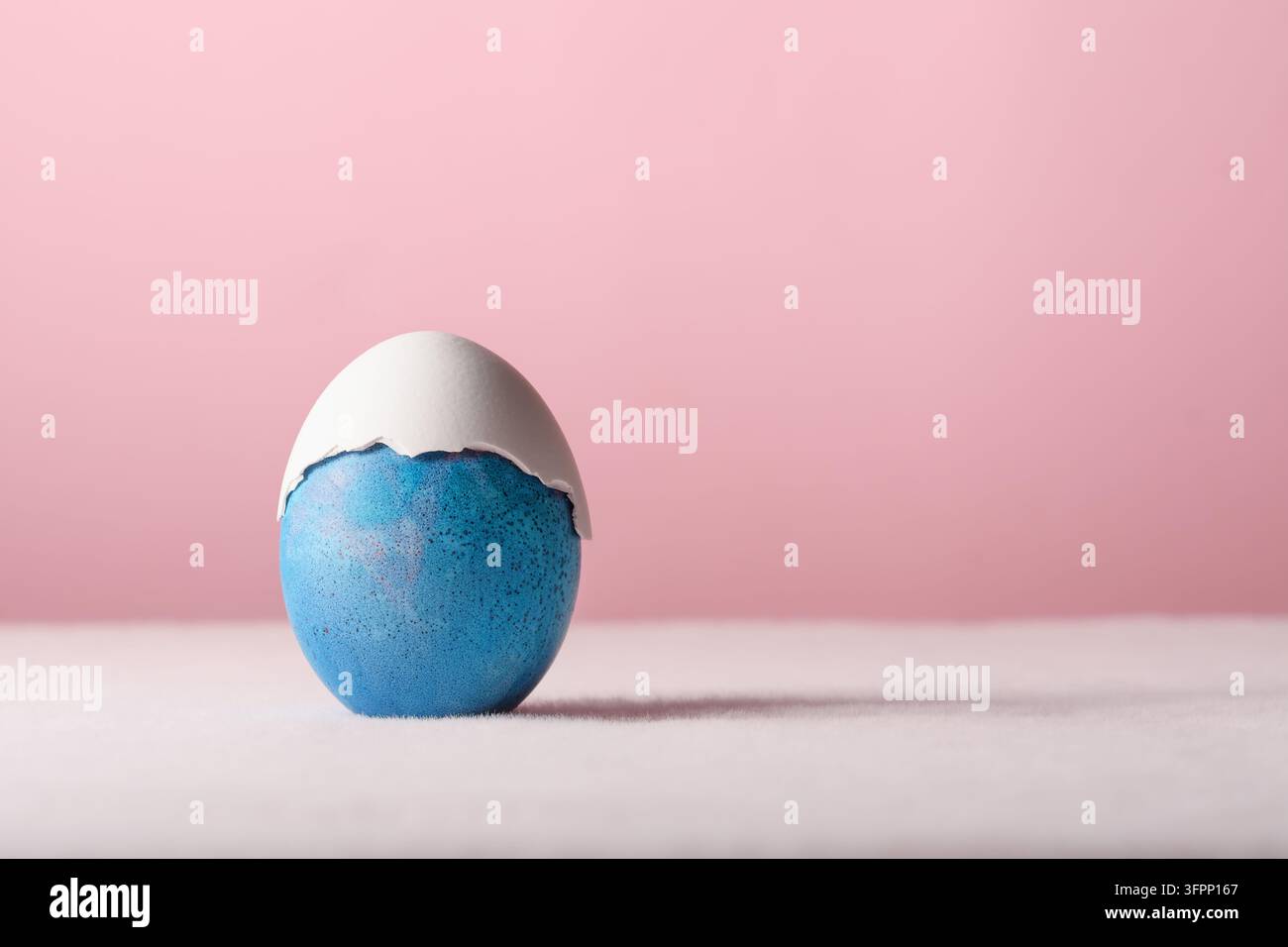Blue Easter egg wears cracked white shell cap, minimalist pastel still life with gentle humor and copy space on pink background. Stock Photo