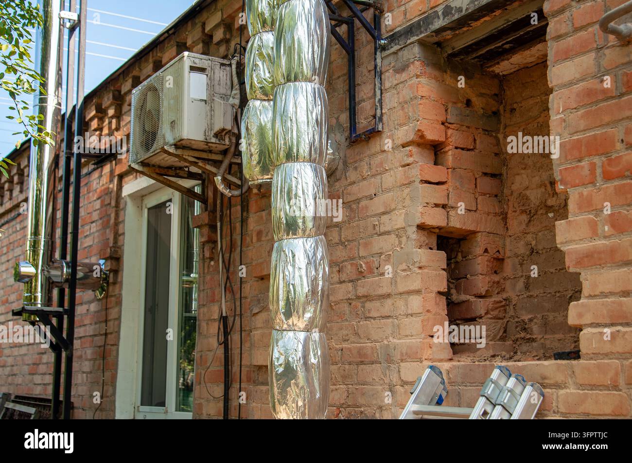 An outdoor air conditioning unit is mounted on a brick wall next to insulated pipes and a window Stock Photo