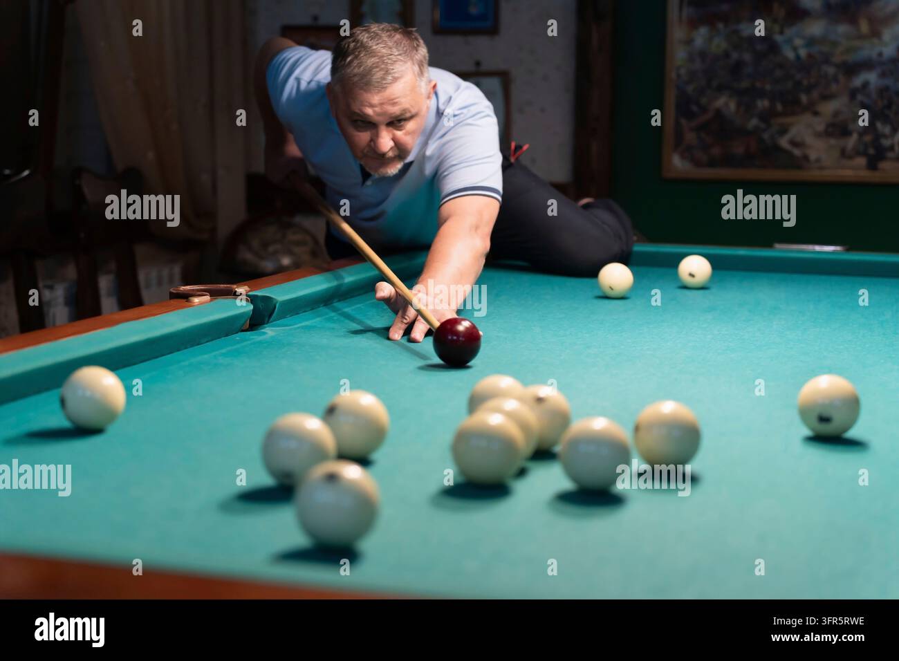 Concentrated mature man playing billiards, carefully aiming his cue stick to meet the white ball on a green pool table in a dimly lit space, showcasin Stock Photo