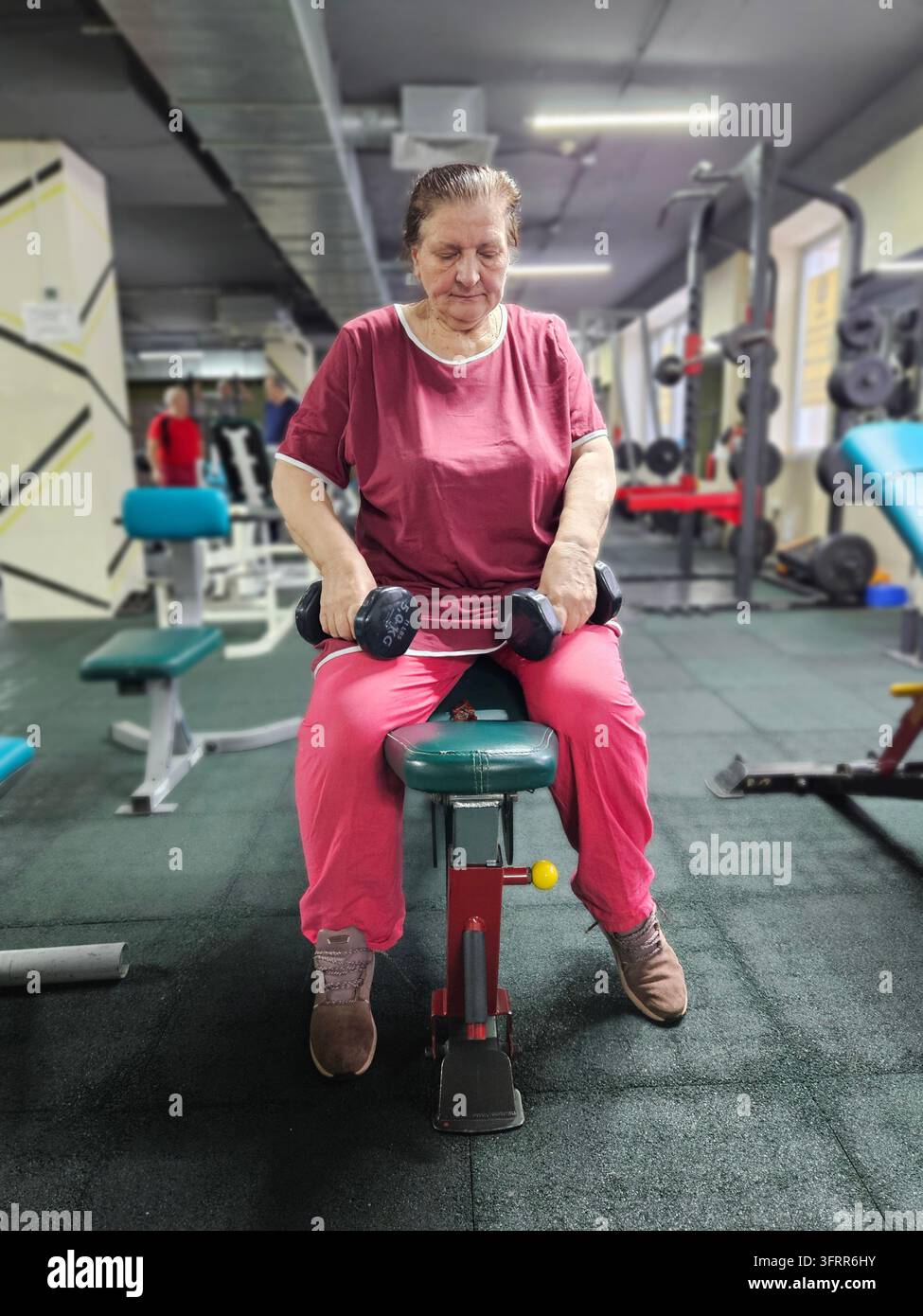 Woman senior in a red shirt and pink pants is holding two dumbbells. She is sitting on a bench Stock Photo