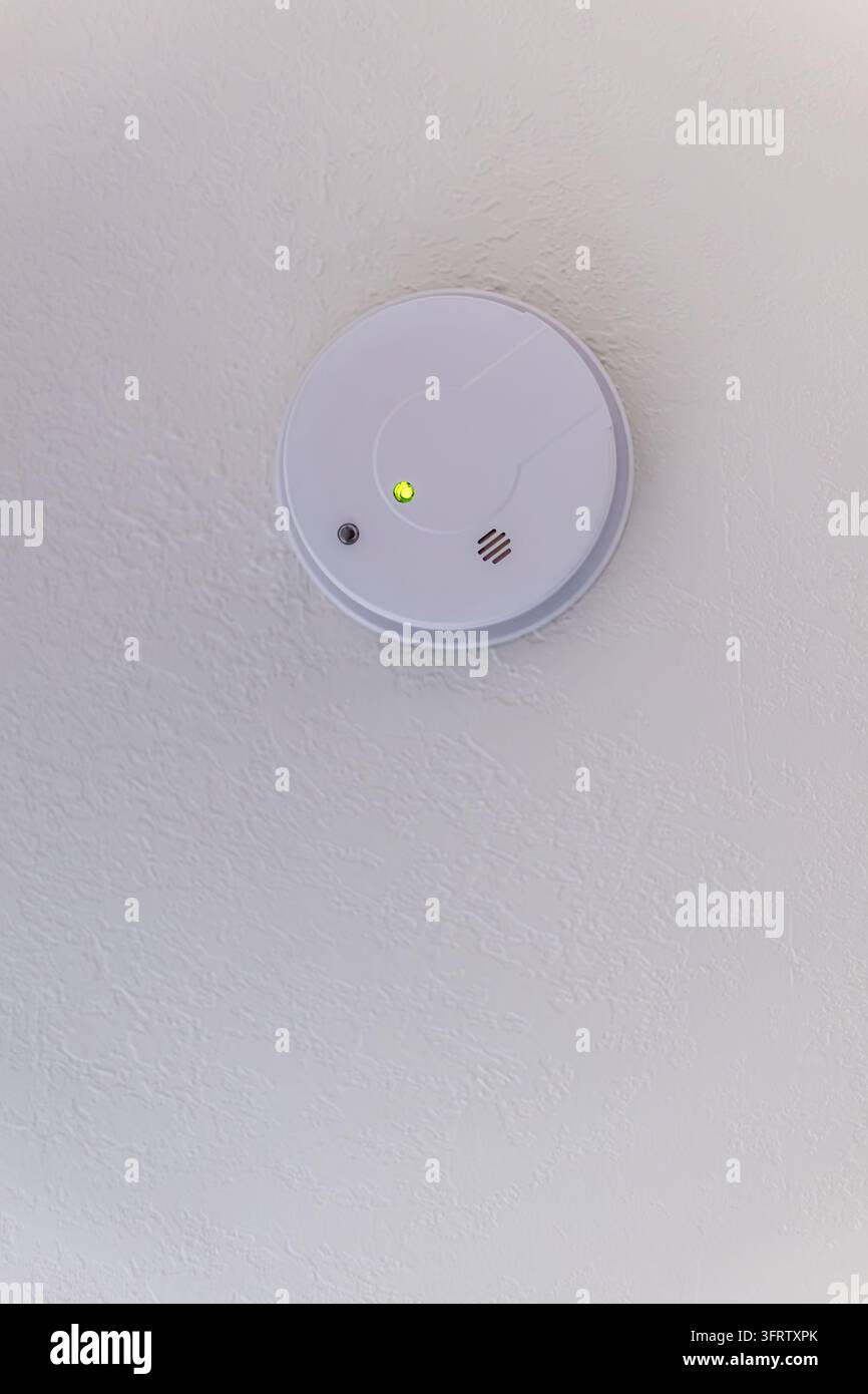 White circular smoke detector mounted on textured wall, featuring glowing green indicator light, compact design and ample negative space. Stock Photo