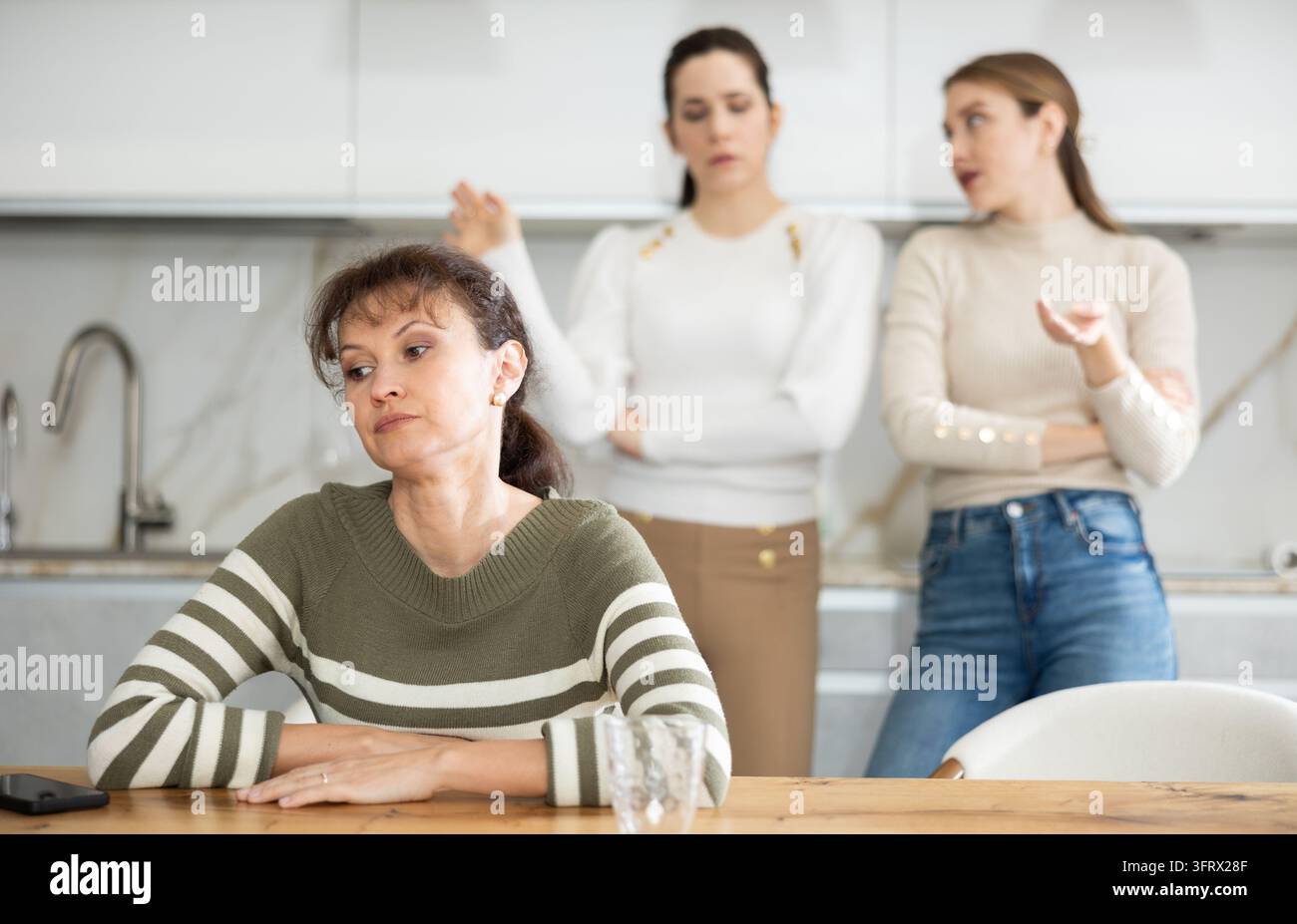 Sad middle-aged woman sitting at table and other women brawling to her ...