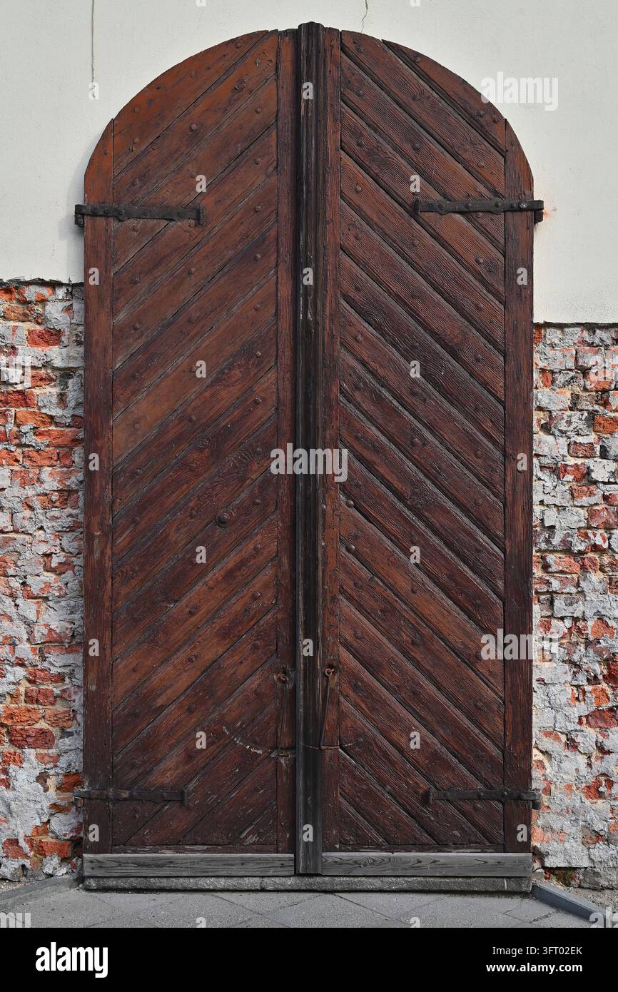 Large arched wooden double door with a herringbone pattern set in a rough brick wall with plaster remnants. Stock Photo