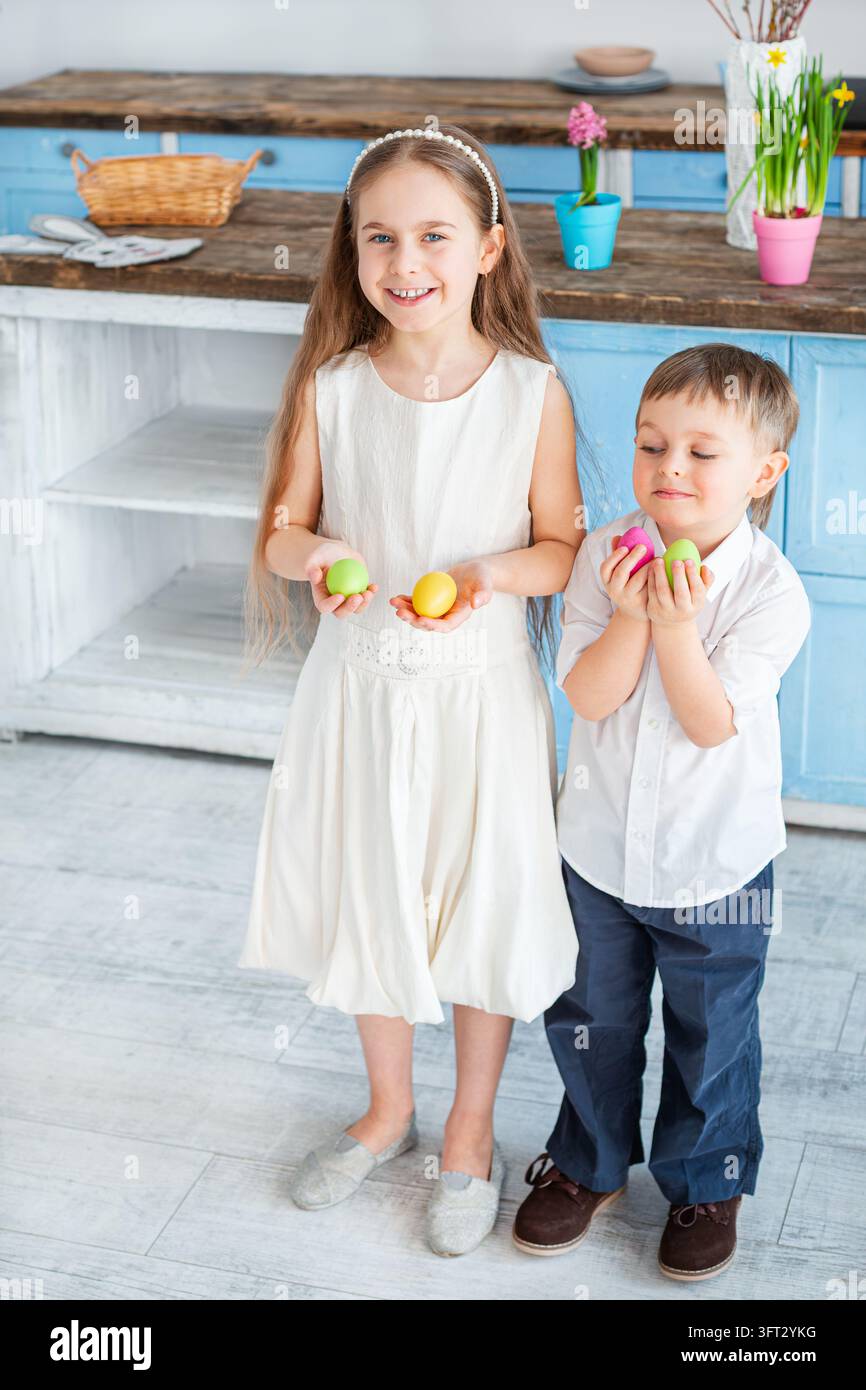 Happy kids holding bright Easter eggs while standing near rustic kitchen counter with flowers and baskets. Stock Photo