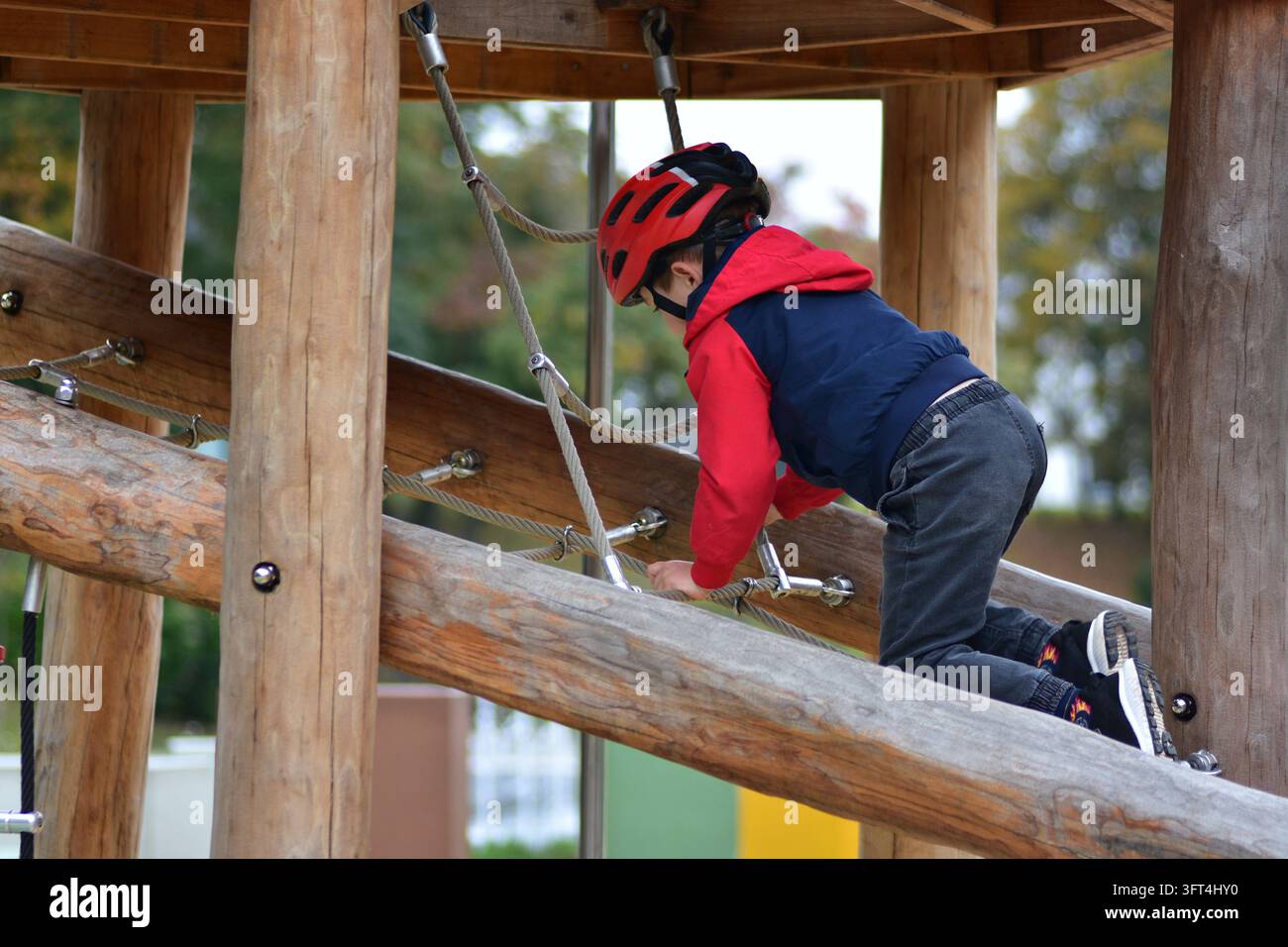 Young boy wearing a red safety helmet climbing on a wooden playground ...
