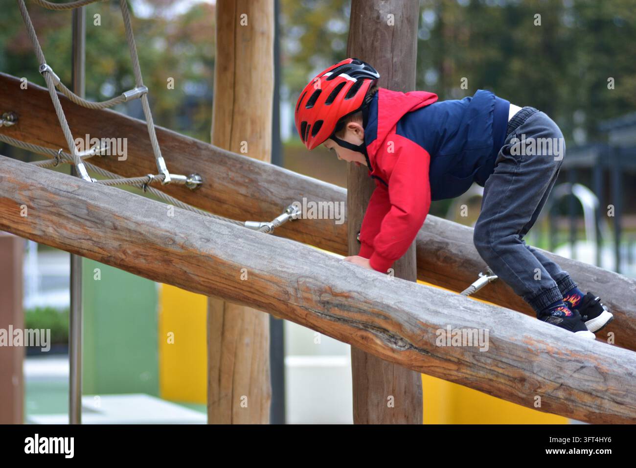 Young boy wearing a red safety helmet climbing on a wooden playground ...