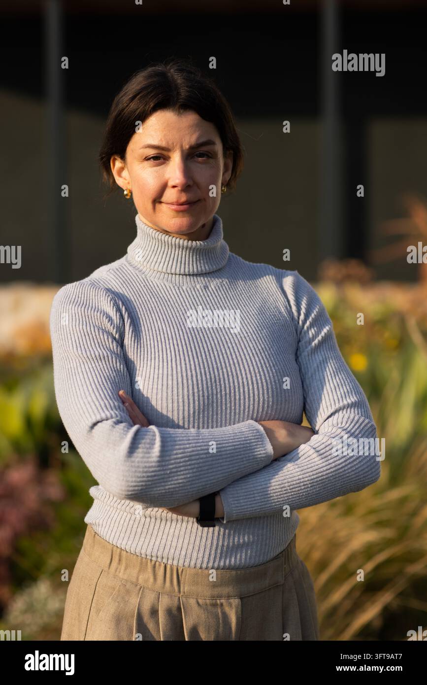 Middle-aged woman standing in courtyard, wearing grey turtleneck with black watch, crossing arms Stock Photo