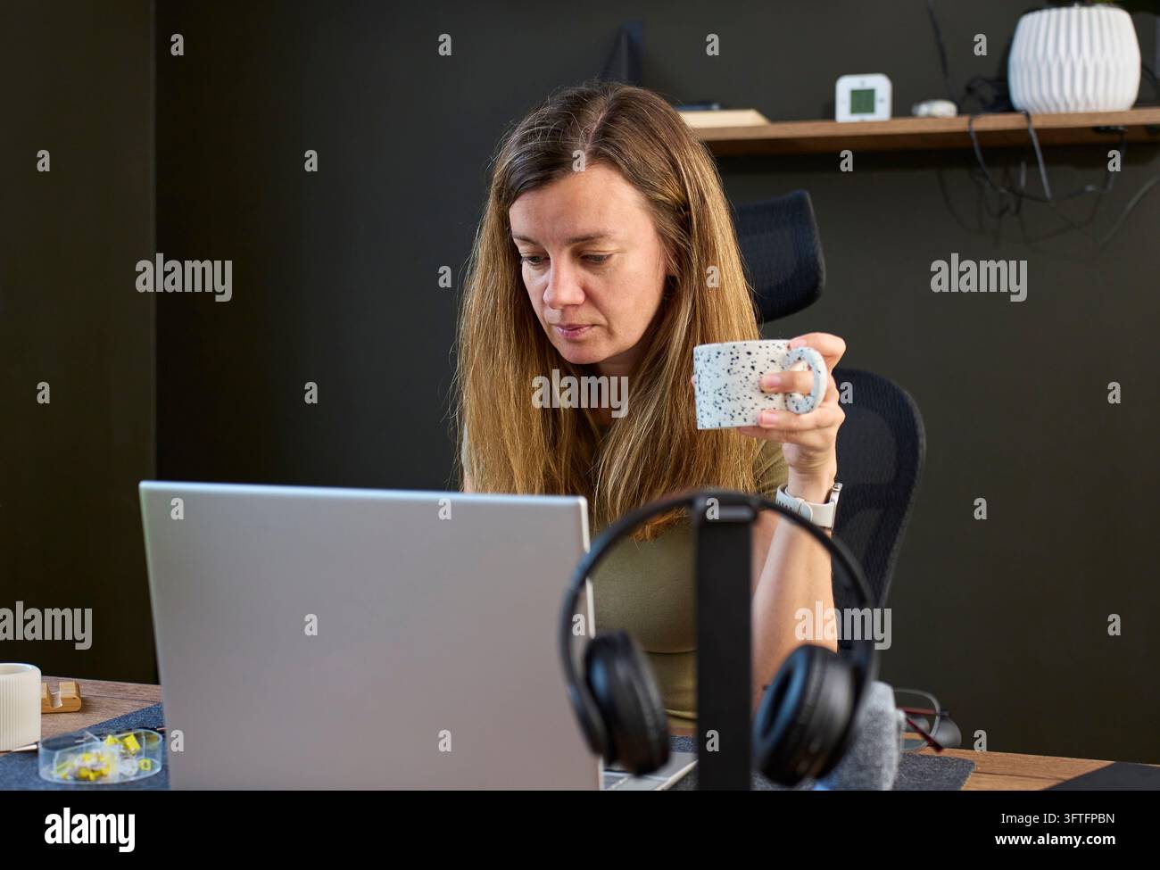 Woman holding coffee mug and looking at laptop screen while sitting at ...