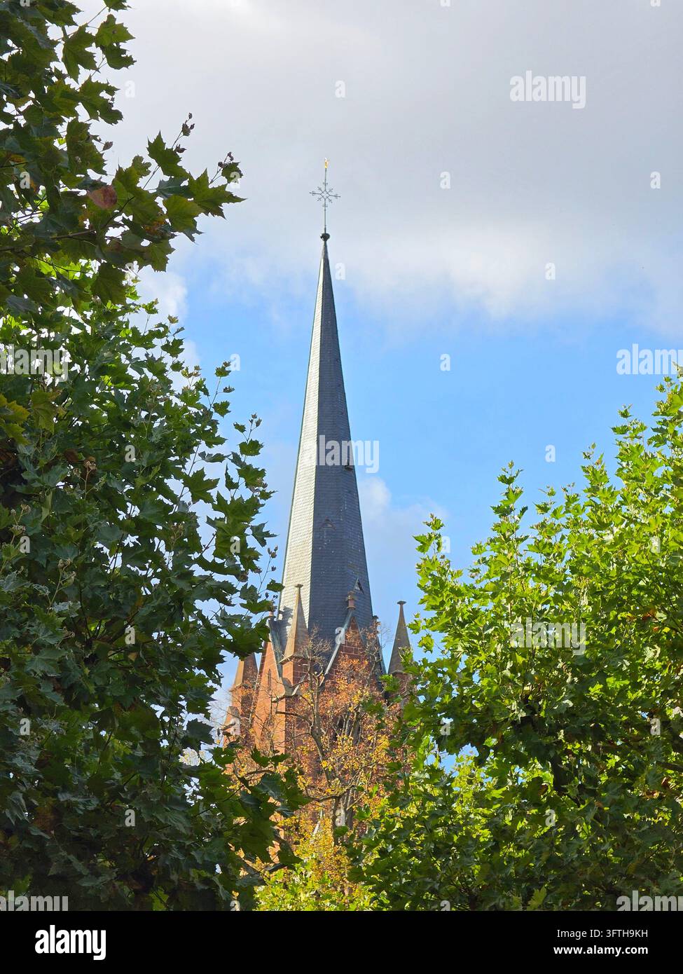 Gothic church spire rising high between green trees under cloudy blue sky vertical portrait landscape orientation view - Smartphone Captured Stock Image