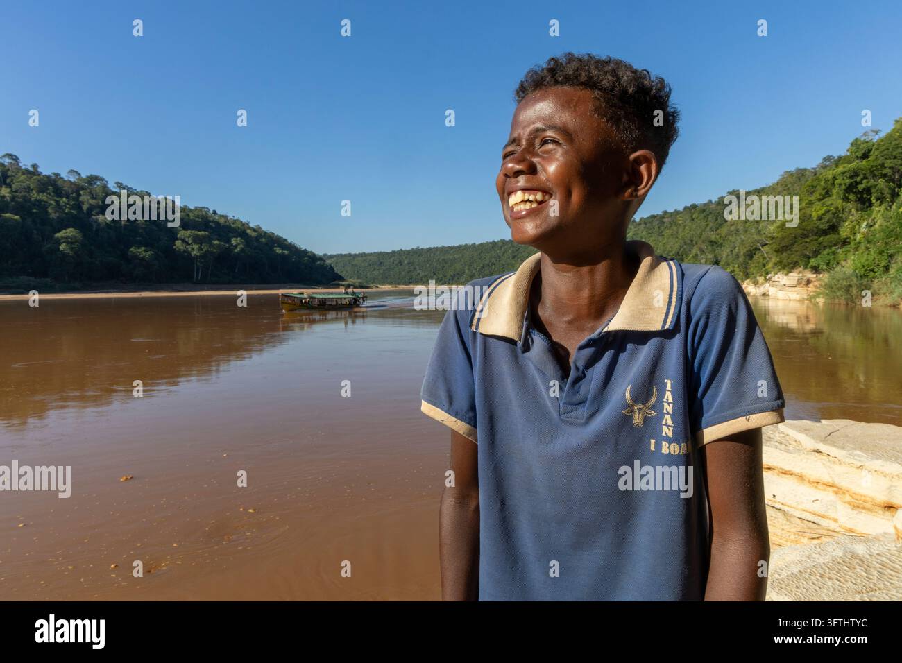 A smiling young man laughs beside the river as a boat sails past ...