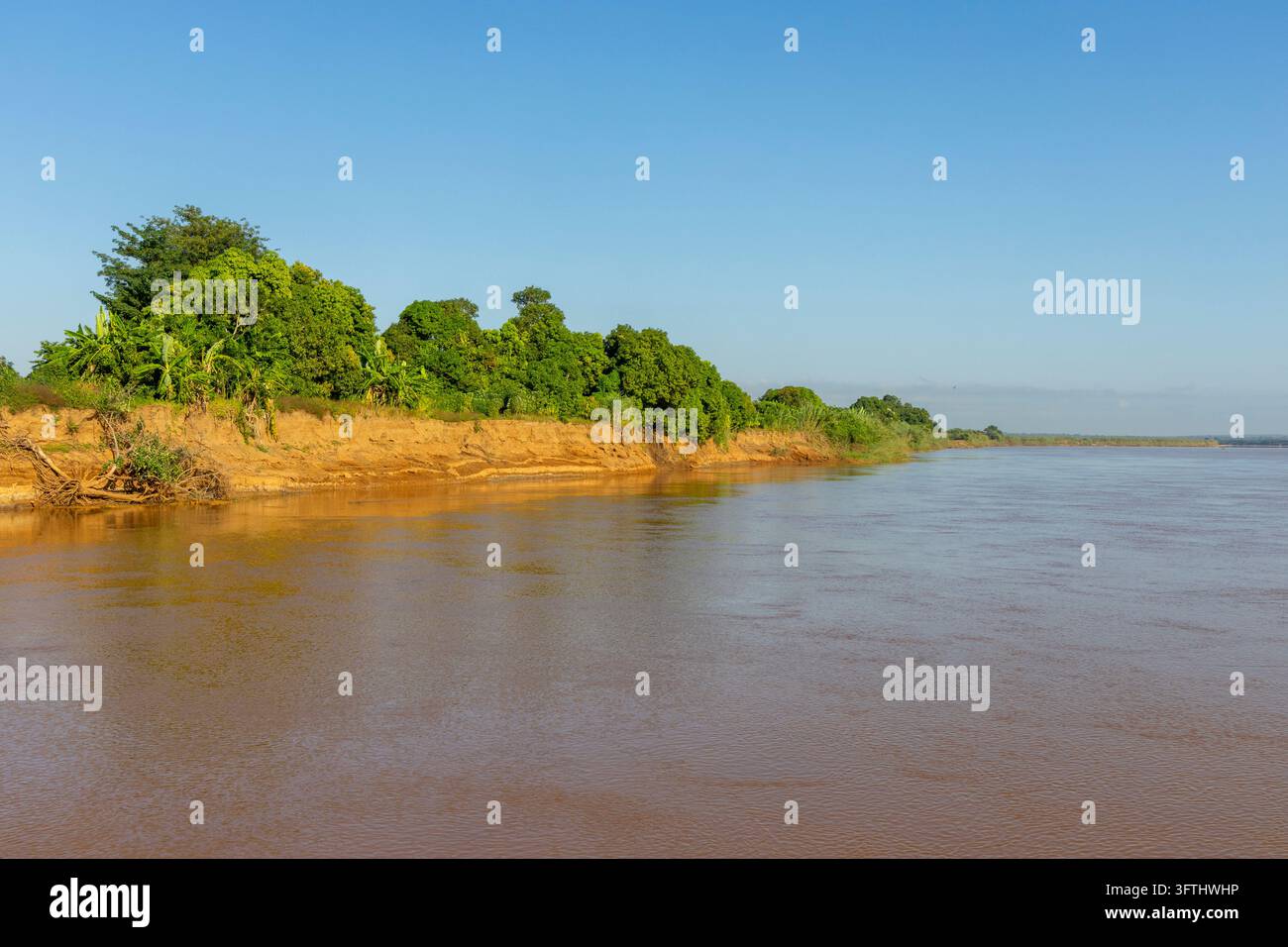 A red eroded riverbank lined with green trees rises beside the calm ...