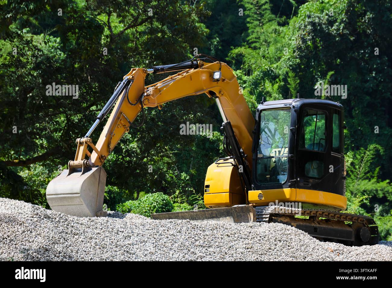 An excavator parked on gravel beside dense green forest, showing yellow ...