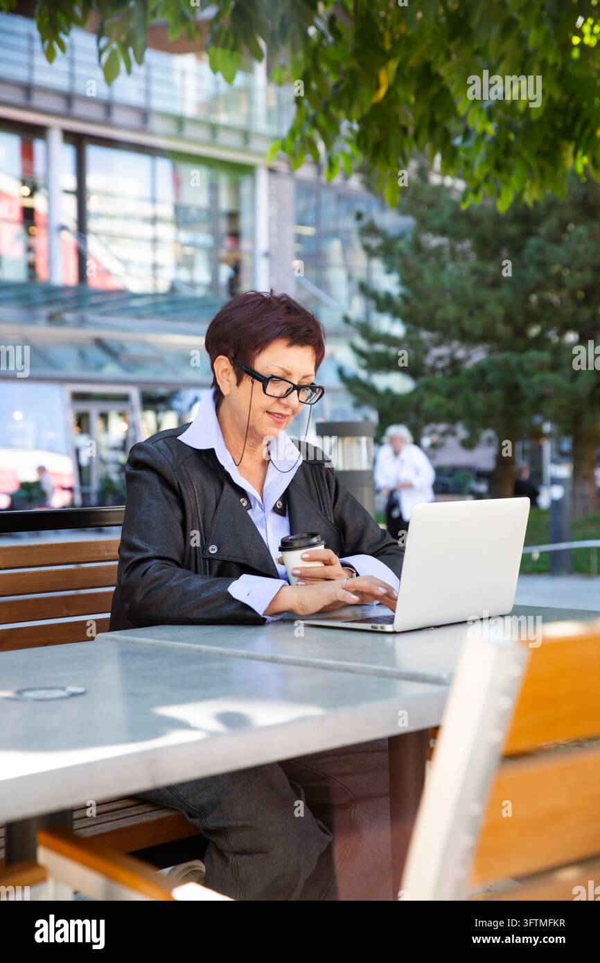 A mature businesswoman works on a laptop with coffee at an outdoor cafe ...