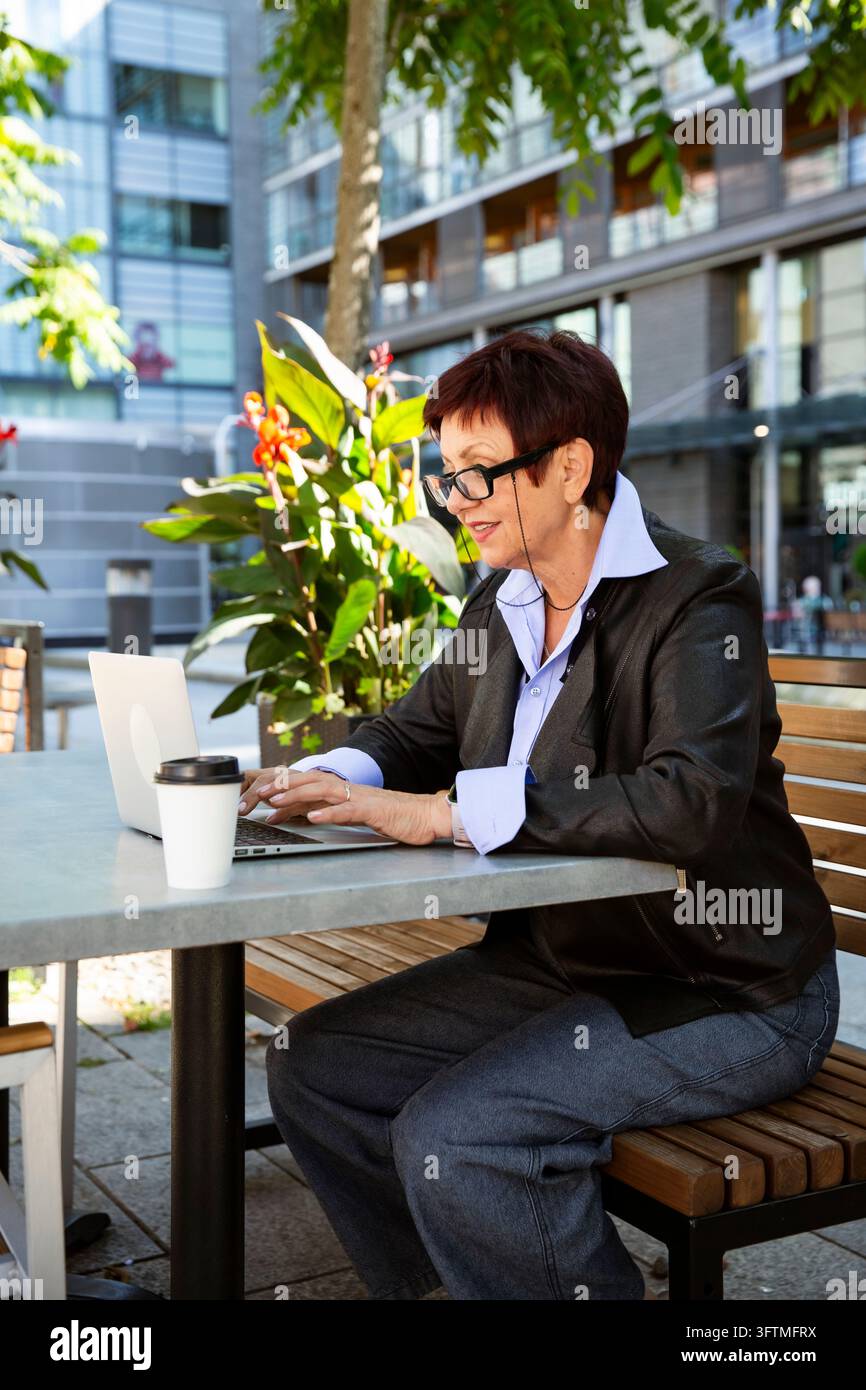 A mature businesswoman works on a laptop with coffee at an outdoor cafe ...