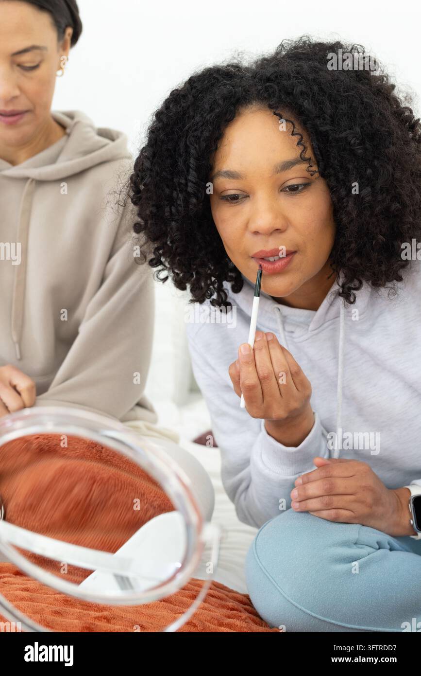 Two female friends wearing hoodies applying lip makeup with brush while ...