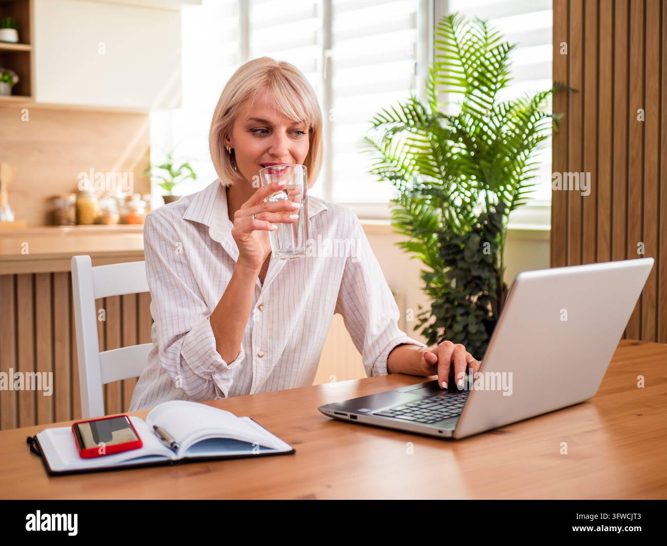Blond woman working from home, typing on laptop and drinking water. Stock Photo