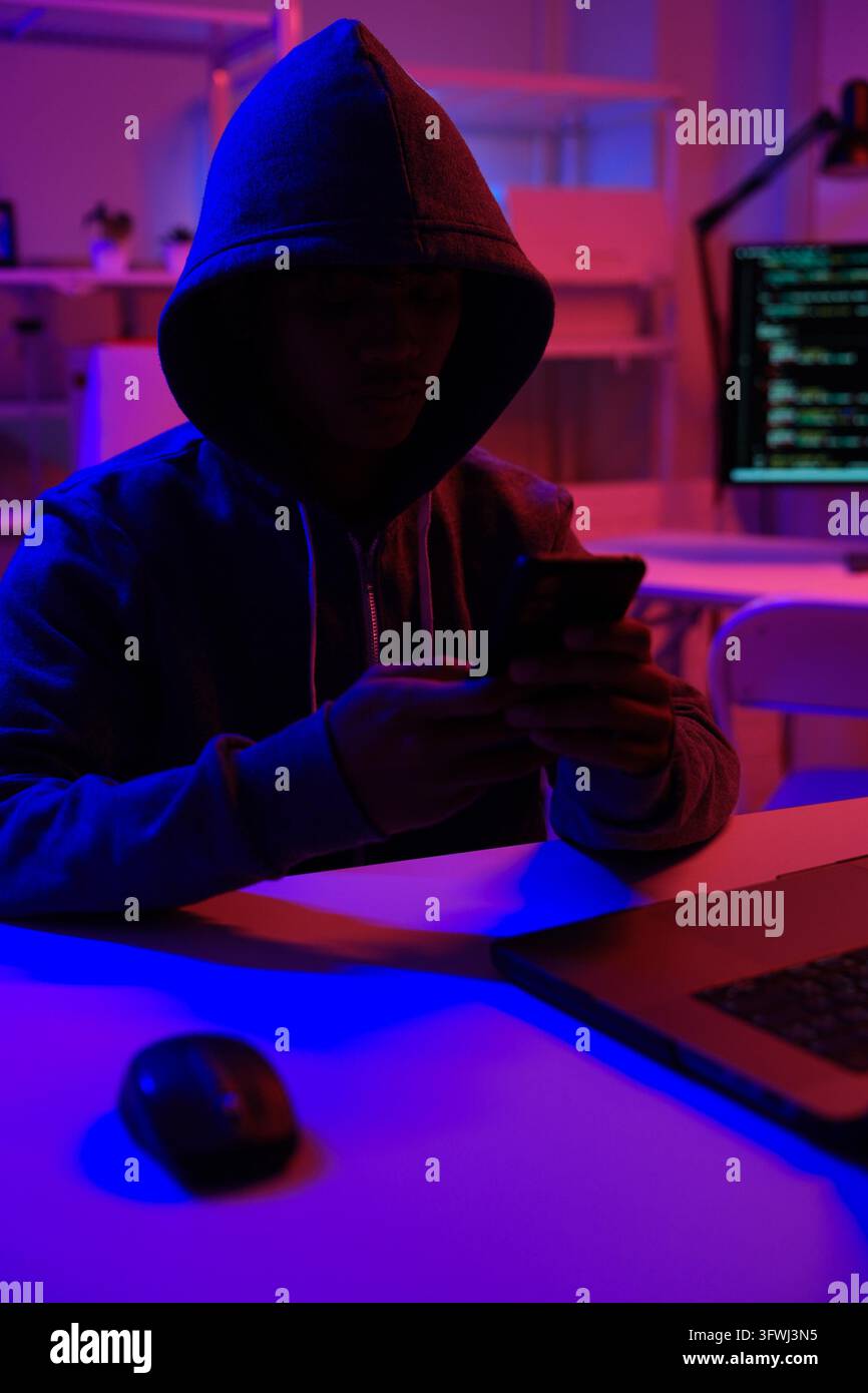 A mysterious hooded hacker working on a computer in a dark room. A concept for cybersecurity, hacking, cybercrime, and data privacy threats. Stock Photo