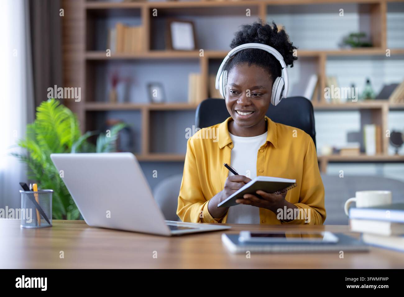 Smiling woman in headphones writes notes beside laptop at office desk ...