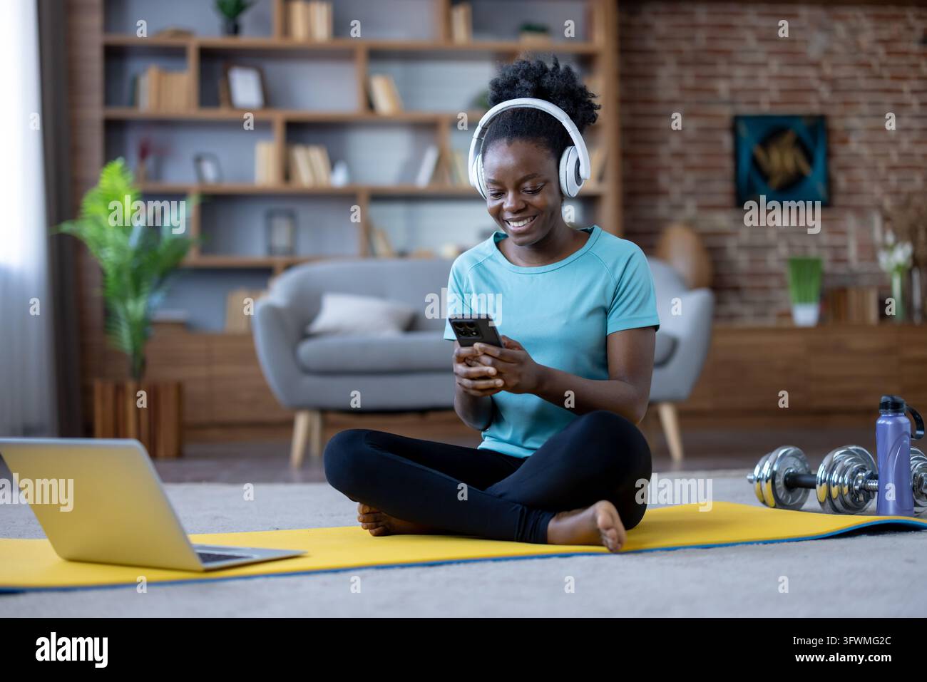 Young woman smiles while using phone during home fitness break, sitting ...