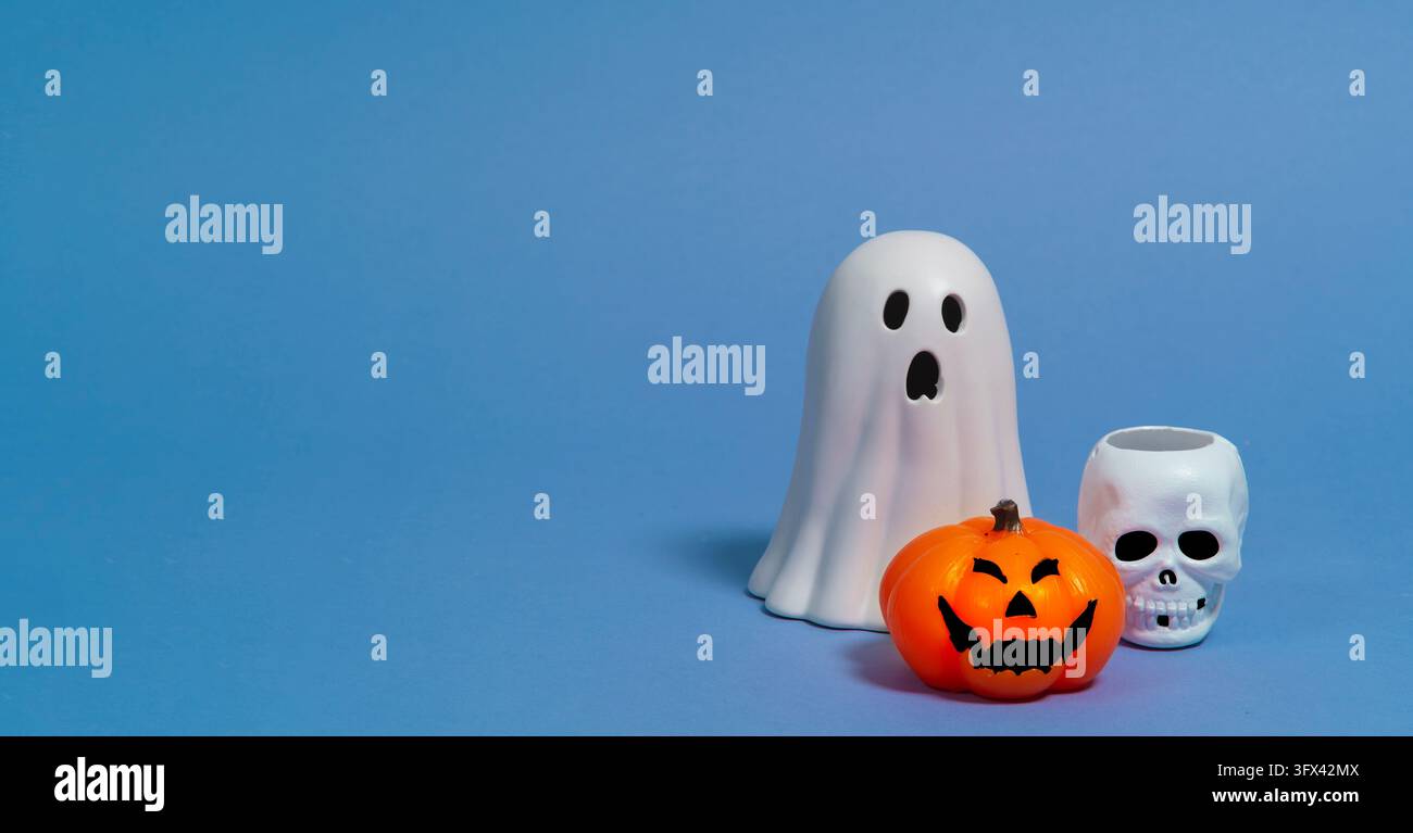 Front view of an orange ceramic pumpkin with a black face, a white skull, and a white ceramic ghost against a blue background Stock Photo