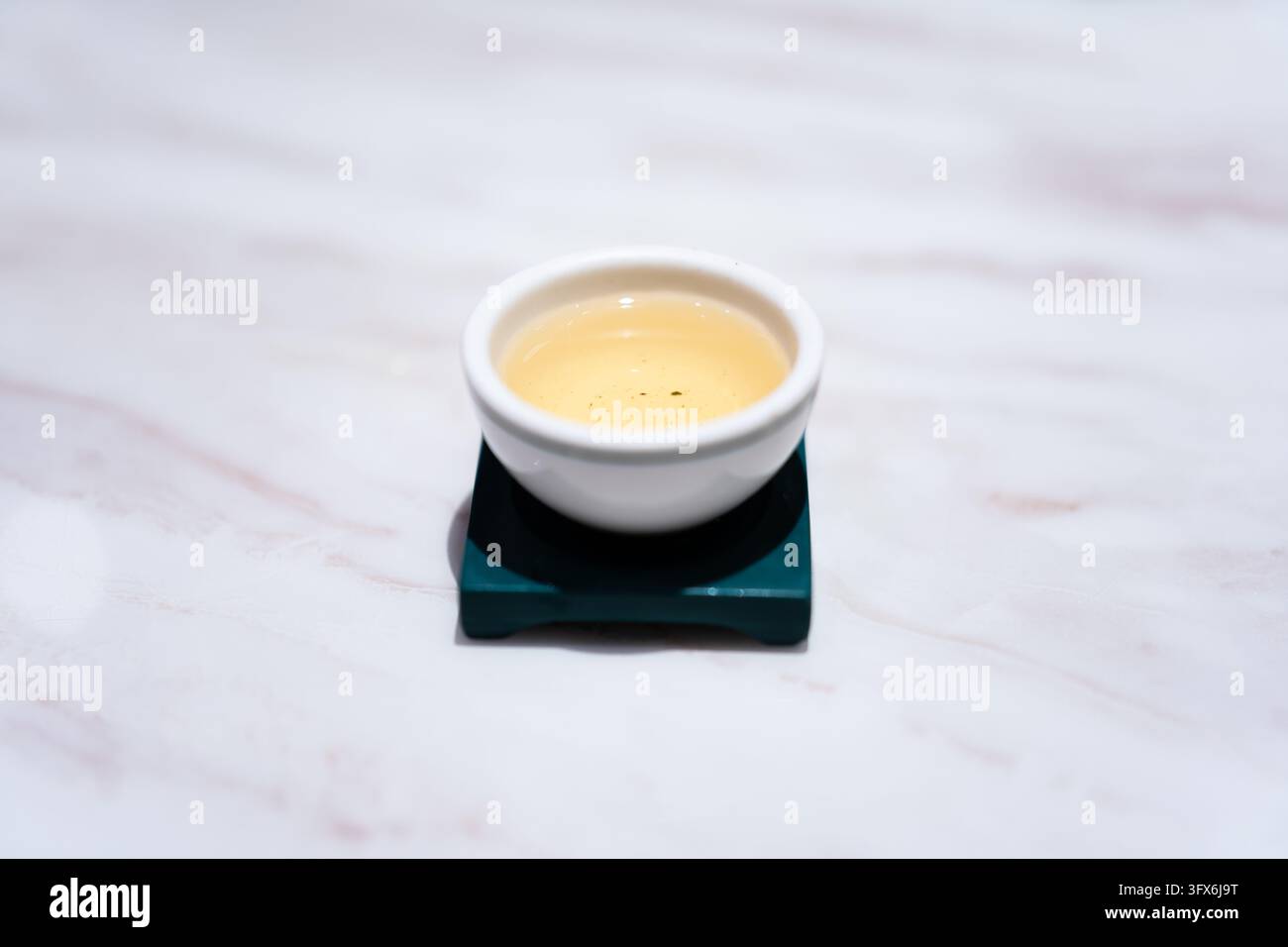 A small white ceramic teacup filled with pale golden oolong tea, resting on a dark green square ceramic saucer, placed on a light veined marble surface in soft diffused lighting, evoking tranquility and traditional East Asian tea culture Stock Photo