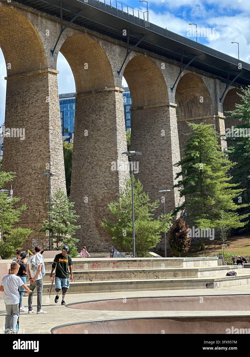 Massive stone viaduct bridge overlooking an urban skatepark with people. Low angle view of a huge historic stone arch viaduct with towering pillars - Smartphone Captured Stock Image