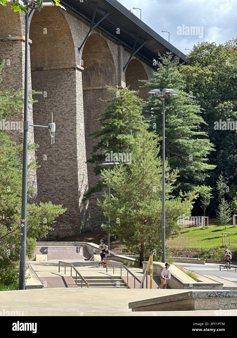 Skate park surrounded by green trees beneath massive historic stone bridge viaduct. Skateboarders active at an outdoor urban skatepark surrounded by l - Smartphone Captured Stock Image