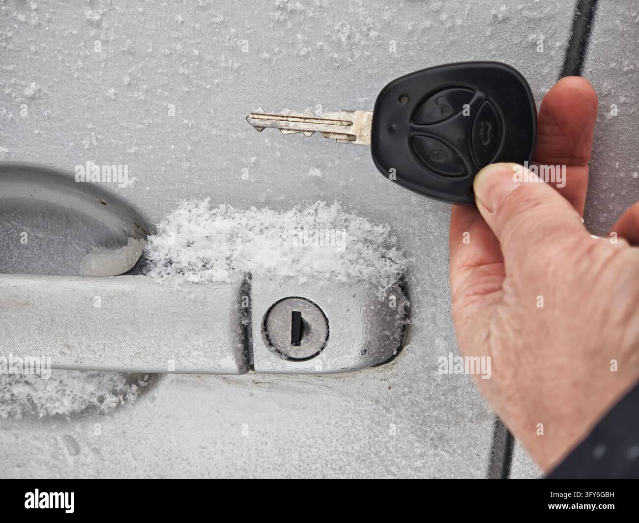 A car door lock is covered with snow and ice while a hand holds a key ...