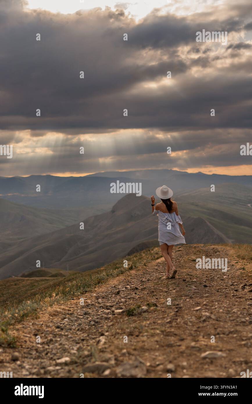 Woman with a hat walks on a rocky hill toward distant mountains under a ...